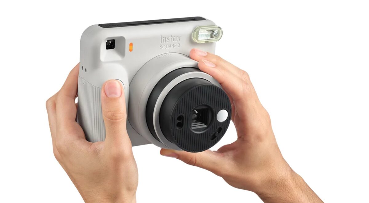 دو دست که یک دوربین چاپ سریع طوسی روشن Instax Square SQ2 را برای عکاسی نگه داشته‌اند، با تمرکز بر کلیدها، لنز چرخشی مشکی و فلاش بالایی در پس‌زمینه سفید.